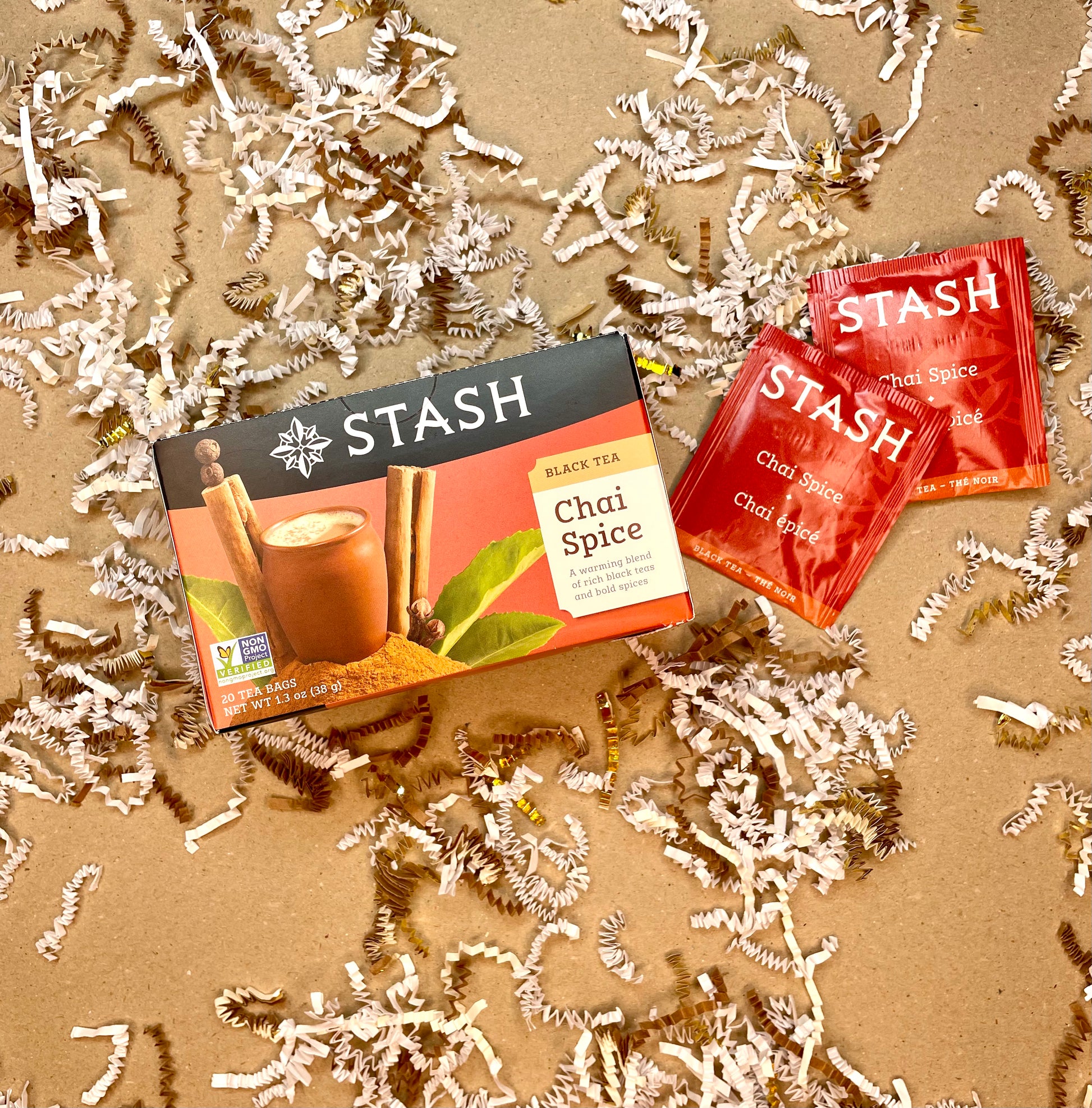 STASH Chai Spice Tea box with 2 individual red tea bags laying next to the box. Backdrop is brown kraft paper color with strips of white, brown, & gold crinkle cut paper.